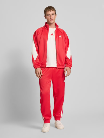 Männermodel trägt Trainingsjacke mit Stehkragen und Logo von adidas Sportswear in Rot mit weißem Logo, Material-Mix, Ganzkörperansicht von vorne.