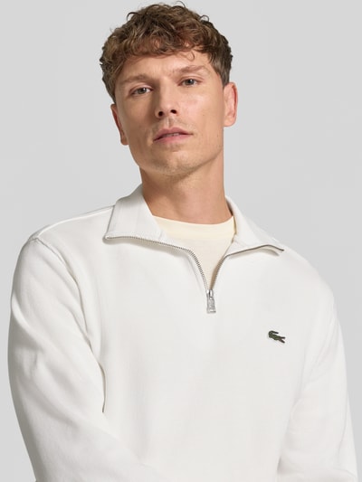 Männermodel trägt Regular Fit Troyer aus reiner Baumwolle von Lacoste in Weiß mit Reißverschluss und Logo, Nahaufnahme Oberkörper. Herren Troyer von Lacoste.