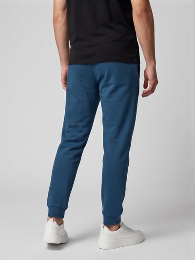 Männermodel trägt blaue Jogginghose, schwarze Shirt, weiße Sneaker, glatter Hintergrund, Ganzkörperansicht Rückansicht.