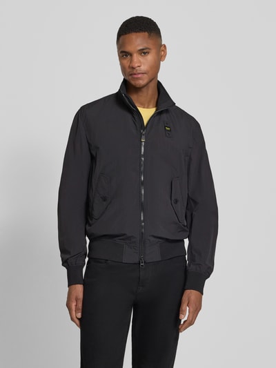 Męski model nosi Jacke mit Pattentaschen Modell 'Alwin' od Blauer Usa, czarna, proste cięcie, stójka, naszywki, przód na suwak, gładka tekstura, widok całej sylwetki z przodu.