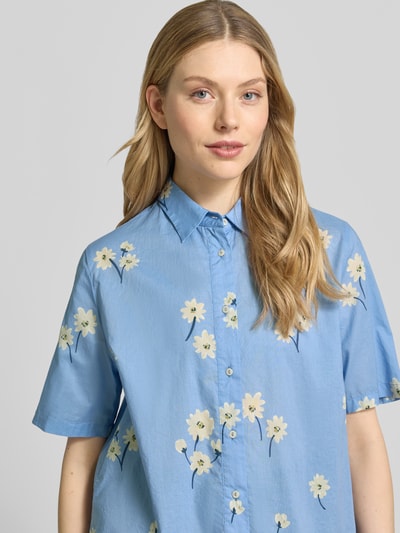 Frauenmodel trägt Relaxed Fit Freizeithemd mit floralem Allover-Print Modell 'Bea' von 0039 Italy aus reiner Baumwolle, hellblau mit gelben Blüten, Kurzärmelig, Kentkragen, durchgehende Knopfleiste, Ganzkörperansicht von vorne.
