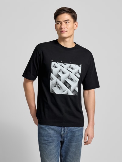 Männermodel trägt Slim Fit T-Shirt aus reiner Baumwolle Modell 'DODGER' von HUGO, Optik: Unifarben, Motiv-Print, Ganzkörperansicht von vorne.