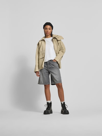 Frauenmodel trägt Jacke aus reiner Baumwolle mit Kapuze von Victoria Beckham, lockere Passform, helle Farbe, seitliche Eingrifftaschen, Reißverschluss und Knopfleiste, Ganzkörperansicht von vorne.