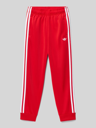 Produktfoto, Trainingshose mit Label-Stitching von adidas Originals in Rot mit weißen Streifen, reguläre Passform, elastischer Bund, Eingrifftaschen, glatte Textur.