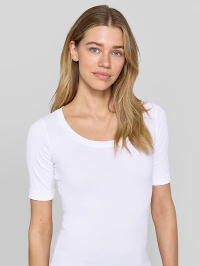 Modelka kobieca ma na sobie T-Shirt mit Ärmelaufschlag und Rundhalsausschnitt od Marc Cain, mieszanka bawełny z dodatkiem streczu, dopasowany krój, okrągły dekolt, rękawy z zakładką, detal z logo, długość tyłu w rozmiarze 36: 60 cm, biały kolor, gładka faktura; zbliżenie na górną część ciała z przodu.