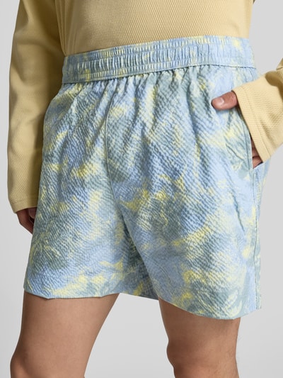 Männermodel trägt Badehose mit Strukturmuster von MAISON KITSUNÉ in Blau-Gelb, Eingrifftaschen, elastischer Bund, Ganzkörperansicht von vorne.