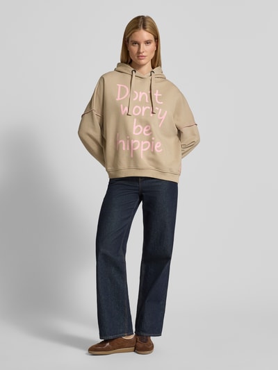 Frauenmodel trägt Oversized Hoodie mit Statement-Print von miss goodlife in Beige mit rosa Schrift, reine Baumwolle, Ganzkörperansicht von vorne.