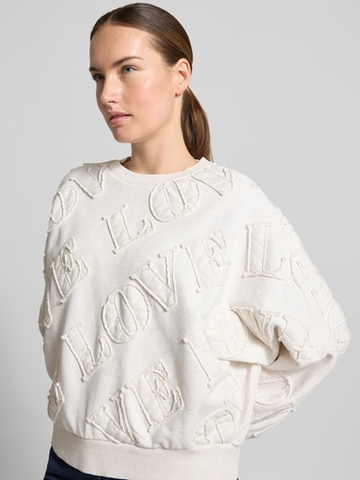 Modelka nosi Oversized Sweatshirt aus reiner Baumwolle Modell 'ASTERA' od DRYKORN, kremowa bluza bawełniana, napisy, wykończenia prążkowane, krój oversized, naszywka z napisem, tekstura boucle, ujęcie z przodu, zbliżenie górnej części ciała.
