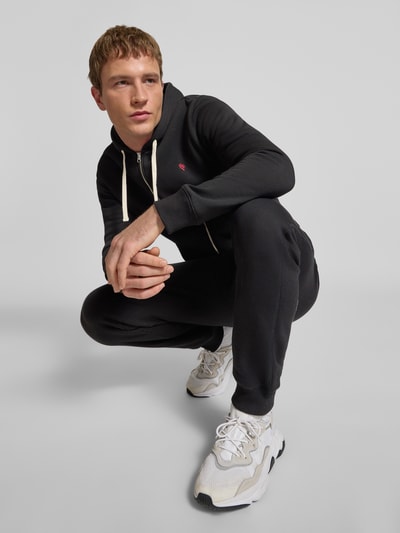 Männermodel trägt Sweatjacke mit Kapuze von REVIEW aus schwarzer Baumwollmischung, Slim-Fit, Reißverschluss, Eingrifftaschen, elastische Abschlüsse, Ganzkörperansicht von vorne.