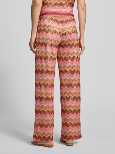 Frauenmodel trägt Wide Fit Stoffhose in Häkel-Optik von Milano Italy in rosa-orangenen Zickzackmustern, semitransparent, mit elastischem Bund, Ganzkörperansicht Rückansicht.