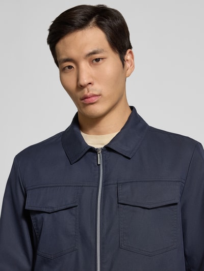 Męski model nosi Jacke mit Pattentaschen und Zweiwege-Reißverschluss od bugatti, bawełniany miks, prosty krój, kent kołnierz, z przodu dwukierunkowy zamek, patkowe kieszenie, boczne kieszenie, długość tyłu w rozmiarze 50: 67 cm, szerokość ramion w rozmiarze 50: 45 cm, szerokość klatki piersiowej w rozmiarze 50: 57 cm, długość rękawa w rozmiarze 50: 70 cm, kolor granatowy, gładka tekstura, zbliżenie na górną część ciała
