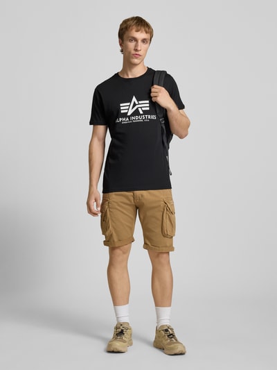 Männermodel trägt T-Shirt mit Logo und Rundhalsausschnitt von Alpha Industries, Schwarz, reine Baumwolle, Regular Fit, Ganzkörperansicht von vorne.