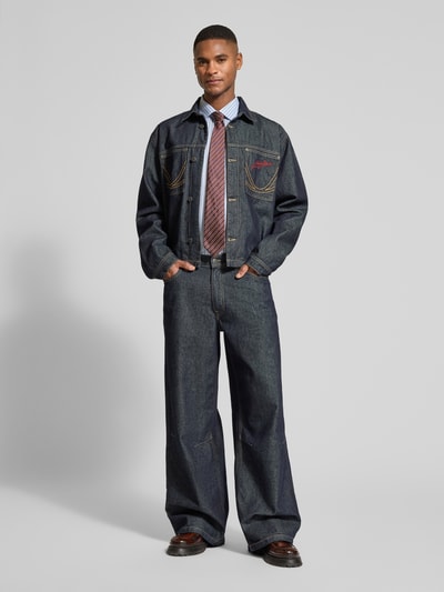 Männermodel trägt Relaxed Fit Jeansjacke mit Ziernähten van KARL KANI, donkerblauw denim met contrasterende stiksels, borstzakken, label-stitching, volledig lichaam vooraanzicht.