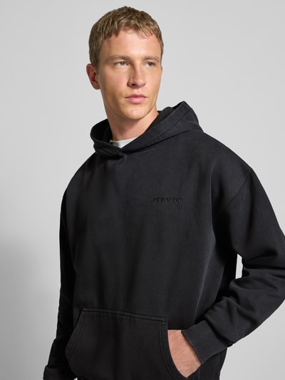 Męski model nosi Oversized Hoodie mit Logo und Känguru-Tasche od Pegador, bawełniany mix, czarny, logo, przód z kieszenią, zbliżenie na górę ciała.