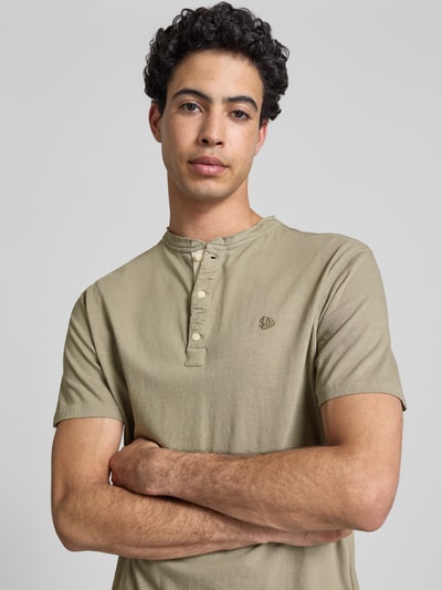 Männermodel trägt T-Shirt mit kurzer Knopfleiste von Superdry in Beige, Baumwoll-Mix mit Leinen-Anteil, Logo-Stitching, Nahaufnahme Oberkörper.