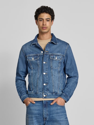 Męski model nosi Regular Fit Jeansjacke aus reiner Baumwolle Modell 'RYAN' von Tommy Jeans, stylizowana na denim, splot twill, naszywka z logo, wytłaczane logo, długi rękaw, wykładany kołnierz, listwa guzikowa, boczne wpuszczane kieszenie i bustasza, pas na plecach, nity; bawełna. Ganzkörperansicht von vorne.