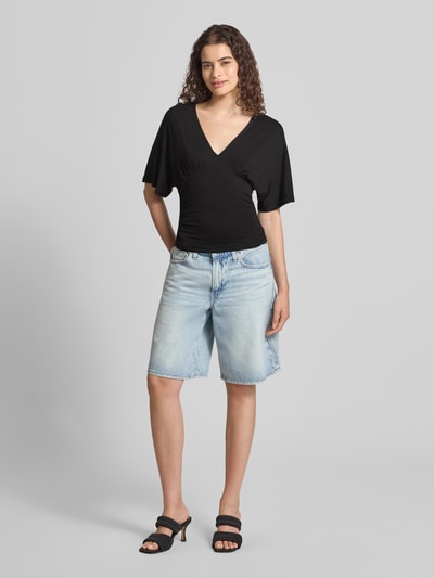 Frauenmodel trägt Bluse aus Viskose-Mix mit V-Ausschnitt von Gina Tricot in Schwarz, taillierte Passform, Halbarm, Ganzkörperansicht von vorne.