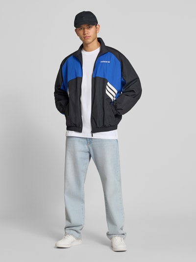 Männermodel trägt Regular Fit Trainingsjacke im Two-Tone-Design Modell 'MUC TT' von adidas Originals in Blau und Schwarz, Ganzkörperansicht von vorne. Herren Trainingsjacke von adidas Originals.