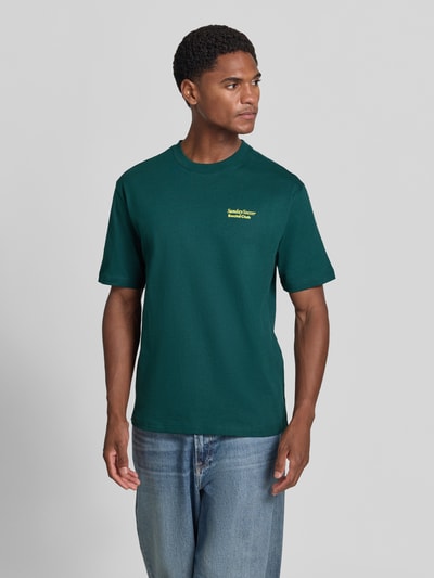 Männermodel trägt T-Shirt mit Statement-Print von MCNEAL in Dunkelgrün mit gelbem Schriftzug, kombiniert mit Jeans, Ganzkörperansicht von vorne, lockerer Schnitt, Rundhalsausschnitt, Rückenlänge bei Größe M 66 cm.