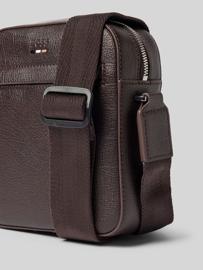 Produktfoto, Umhängetasche mit Label-Detail von BOSS in Dunkelbraun mit grober Leder-Textur und strukturiertem Riemen vor grauem Hintergrund.