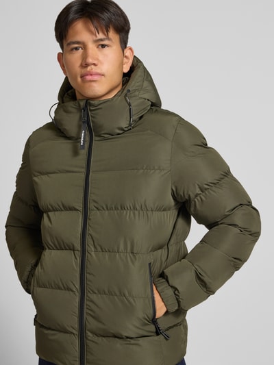 Männermodel trägt Steppjacke mit Label-Stitching und Kapuze von Superdry in Oliv, sichtbarer Reißverschluss, seitliche Eingrifftaschen, Nahaufnahme Oberkörper.