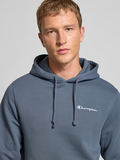 Męski model nosi Hoodie mit Kapuze od CHAMPION z mieszanki bawełny, luźny krój, kaptur z tunelem, wyhaftowane logo, kieszeń kangurka, prążkowane wykończenia, kolor szaroniebieski, zbliżenie na górną część ciała.