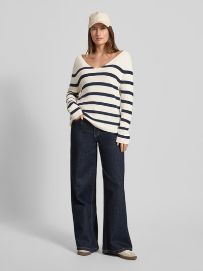 Frauenmodel trägt Regular Fit Strickpullover aus reiner Baumwolle von Tommy Hilfiger, Optik: Allover-Muster, gestreift, Logo-Stitching, V-Ausschnitt, Ärmelform: Langarm mit Raglanärmel, Materialverarbeitung: Rippstrick, Farbe creme mit dunkelblauen Streifen, Ganzkörperansicht von vorne.