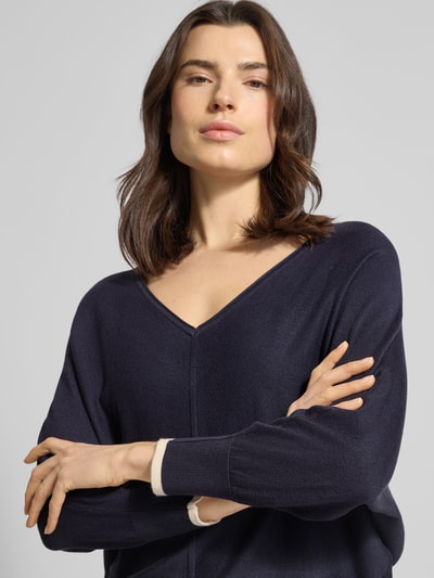 Kobiece modelka nosi Strickpullover aus Viskose-Mix mit V-Ausschnitt od comma, ciemnogranatowy, paski w kontrastowym kolorze na rękawach, krój regular fit, mieszanka wiskozy, dekolt w serek, długość tyłu w rozmiarze S: 52 cm, długość rękawów w rozmiarze S: 56 cm, zbliżenie na górną część ciała z przodu.