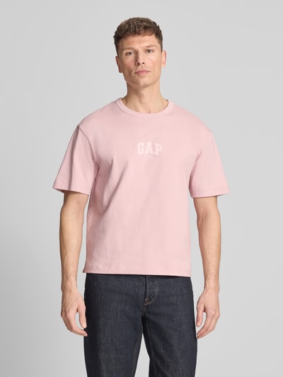 Męski model nosi T-Shirt mit Logo und Rundhalsausschnitt von GAP, różowy, bawełna, krój regular fit, logo, okrągły dekolt, długość tyłu w rozmiarze M: 61 cm, widok całej sylwetki od przodu.