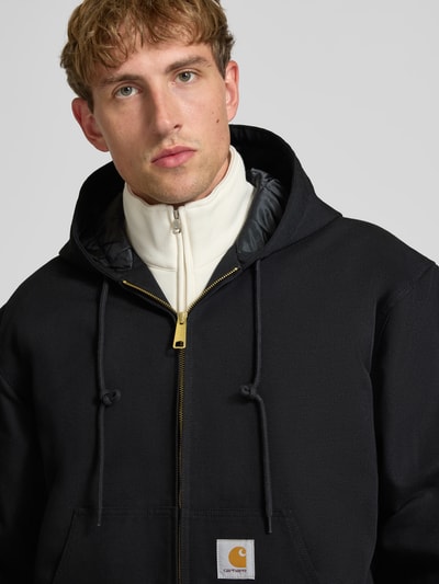 Männermodel trägt Jacke mit Label-Patch und Känguru-Tasche von Carhartt Work In Progress in Schwarz mit Reißverschluss, Nahaufnahme Oberkörper.