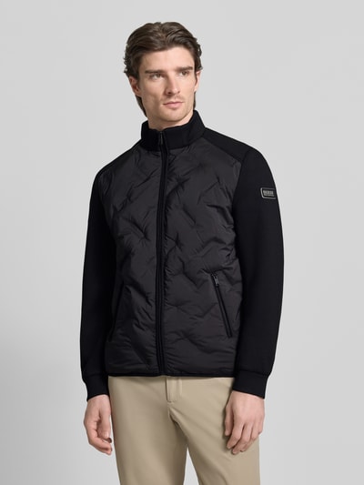Männermodel trägt Regular Fit Jacke mit Stehkragen von Guess, schwarz, Reißverschluss, Eingrifftaschen, elastischer Material-Mix, Ganzkörperansicht von vorne.