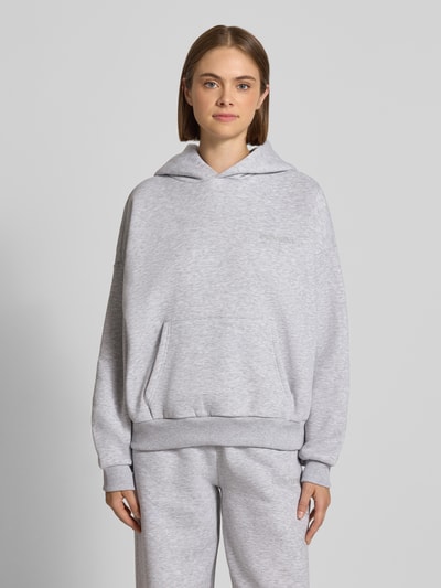Modelka nosi Relaxed Fit Basic Hoodie in grau melange od REVIEW, mieszanka bawełny, krój relaxed fit, kaptur, nadruk z logo, kieszeń kangurka, prążkowane wykończenia, widok całej sylwetki z przodu.
