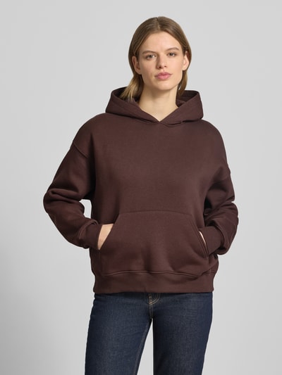 Modelka kobieca nosi Regular Fit Hoodie mit Känguru-Tasche Modell 'Iconic' od Gina Tricot, mieszanka bawełny, krój regular fit, kieszeń kangurka, elastyczne wykończenia, ciemnobrązowy kolor, gładka tekstura, widok na całą sylwetkę z przodu.