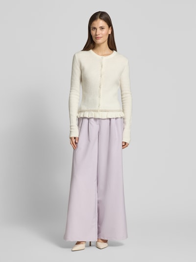 Frauenmodel trägt Slim Fit Cardigan in Strick-Optik Modell 'Kate' von GUIDO MARIA KRETSCHMER WOMAN mit Rüschen- und Ziersteinbesatz, cremefarben, kombiniert mit weiten fliederfarbenen Hosen, feine Textur, Ganzkörperansicht von vorne.