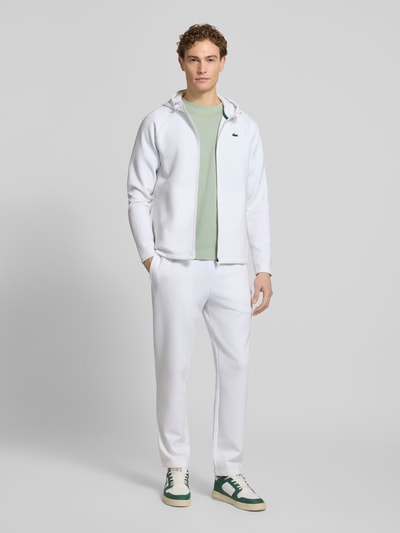 Männermodel trägt Regular Fit Sweatjacke mit Baumwoll-Anteil von Lacoste, Optik: Unifarben, Passform: Regular-Fit, Materialverarbeitung: Scuba, Material: Baumwoll-Mix, mit Kapuze, Reißverschluss, Logo-Patch, seitliche Eingrifftaschen, Farbe Weiß, Ganzkörperansicht von vorne.