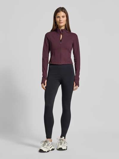 Kobiece modelka nosi kurtkę Slim Fit Jacke mit Stehkragen Modell 'Storm' od Fabletics w kolorze ciemnofioletowym, gładka faktura, pętelki na kciuki, krój slim fit, krótki krój, Szerokość ramion w rozmiarze S: 36 cm, Szerokość klatki piersiowej w rozmiarze S: 42 cm, Długość tyłu w rozmiarze S: 44 cm, Długość rękawów w rozmiarze S: 74 cm, widok całej sylwetki z przodu.