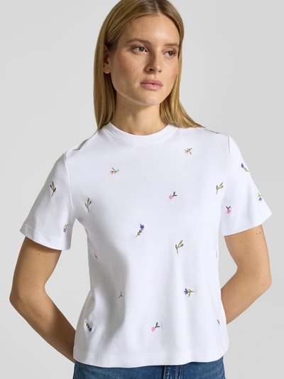 Modelka kobieca nosi T-Shirt mit Blumenstickerei von Christian Berg Woman z białej bawełny, prosty krój, haftowane kwiaty, dekolt okrągły, półrękaw, widok na górną część ciała z przodu.
