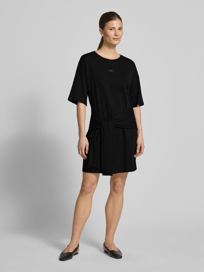 Modelka kobieca nosi T-Shirt-Kleid mit Rundhalsausschnitt und 1/2-Arm od ARMANI EXCHANGE, czarna bawełna, prosty krój, minikleid, detal z logo, długość tyłu S: 82 cm, szerokość w rozmiarze S: 50 cm, długość rękawa S: 31 cm, całopostaciowy widok z przodu.