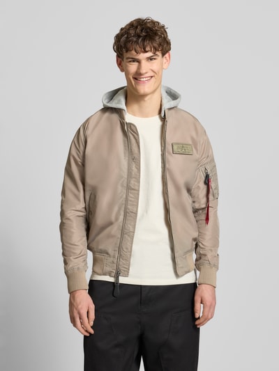 Männermodel trägt Blouson mit Logo-Anhänger Modell 'MA-1 TT' von Alpha Industries in Beige mit Sweat-Kapuze, Label-Patch, Nahaufnahme Oberkörper. Lockere Passform, elastische Abschlüsse, Armtasche, Reißverschluss sichtbar.