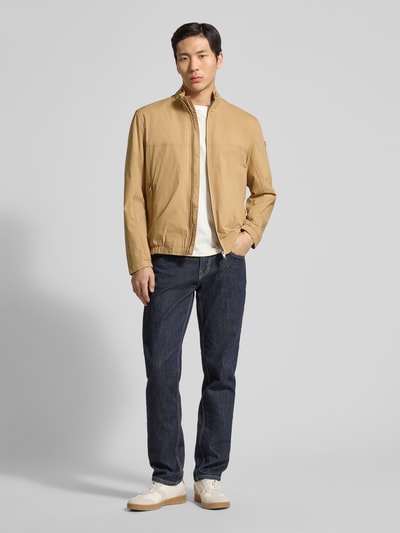 Männermodel trägt heren jack model 'Calvin' van Brax in katoenmix met stretch, recht model, opstaande kraag, ritszakken, ritssluiting, beige tint, jeans en sneakers. Ganzkörperansicht von vorne.