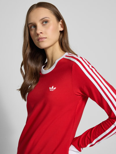 Frauenmodel trägt Longsleeve mit Rundhalsausschnitt und Logo-Stitching von adidas Originals, Baumwoll-Mix mit Stretch-Anteil, körpernaher Schnitt, Rot mit weißen Details, Rückenlänge bei Größe S 53 cm, Nahaufnahme Oberkörper.