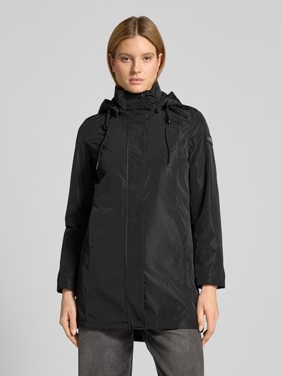 Frauenmodel trägt Regular Fit Mantel mit Logo-Applikation von NAUTGÅRD in Schwarz, mit Stehkragen, Kapuze mit Tunnelzug, Eingrifftaschen, Ganzkörperansicht von vorne. Rückenlänge 88 cm, Schulterbreite 37 cm, Brustweite 48 cm, Ärmellänge 63 cm bei Größe S.