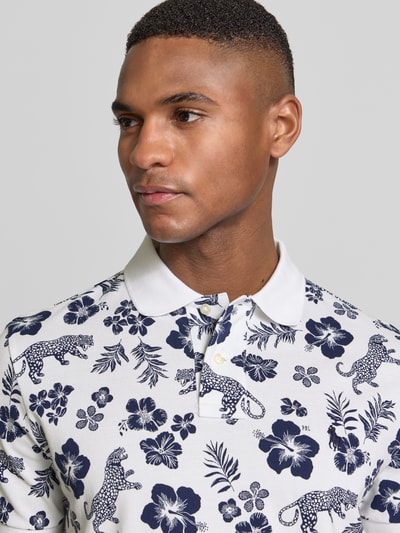 Męski model nosi Slim Fit Poloshirt aus reiner Baumwolle von Polo Ralph Lauren, biało-granatowy wzór na całej powierzchni, detal z logo, kołnierzyk polo, dekolt w serek, krótki rękaw, prążkowane zakończenia, boczne rozcięcia, wykończenie materiału: pika, czysta bawełna. Zbliżenie górnej części ciała.