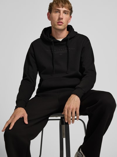 Männermodel trägt Relaxed Fit Hoodie aus Baumwoll-Mix mit Label-Detail von Alpha Industries, schwarz, Nahaufnahme Oberkörper.