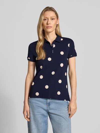 Modelka nosi koszulkę polo z krótką listwą guzikową od Christian Berg Woman, mieszanka bawełny i elastanu, dopasowany krój, kołnierzyk polo, długość tyłu 61 cm w rozmiarze S, granatowe tło z jasnymi groszkami, gładka tekstura. Ujęcie: całkowita sylwetka z przodu.