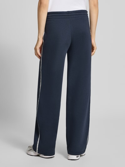 Kobieta modelka ma na sobie ciemnoniebieskie spodnie dresowe Wide Fit Sweatpants aus Baumwoll-Mix od REVIEW, mieszanka bawełny, krój wide fit, elastyczny pas, bordiura, wyhaftowane logo, długość wewnętrzna nogawki w rozmiarze S 44 cm, widok całej sylwetki od tyłu.