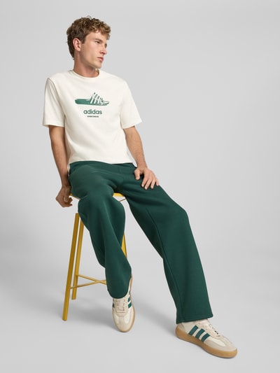 Männermodel trägt Regular Fit T-Shirt aus reiner Baumwolle Modell 'SLIDE' von adidas Sportswear w kolorze ecru z zielonym nadrukiem, prosta forma, gładka tekstura, Ganzkörperansicht von vorne