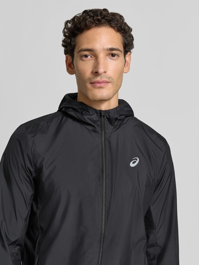 Männermodel trägt schwarze Regular Fit Trainingsjacke mit Logo-Print Modell 'Core' von ASICS TIGER, glatte Textur, Nahaufnahme Oberkörper.