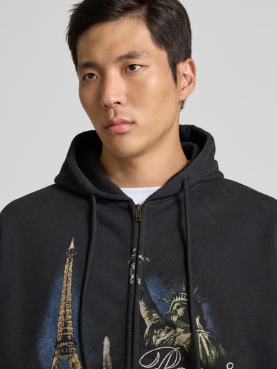 Männermodel trägt Oversized Sweatjacke mit Foto-Print von VETEMENTS in Schwarz, Zweiwege-Reißverschluss, Baumwoll-Mix, Nahaufnahme Oberkörper. Rückenlänge 74 cm, Schulterbreite 68 cm, Ärmellänge 67 cm, Oversized-Fit.