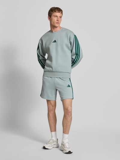 Männermodel trägt Regular Fit Shorts mit Viskose-Anteil von adidas Sportswear im Baumwoll-Viskose-Mix, Logo-Stitching, Farbton Hellblau, Ganzkörperansicht von vorne.